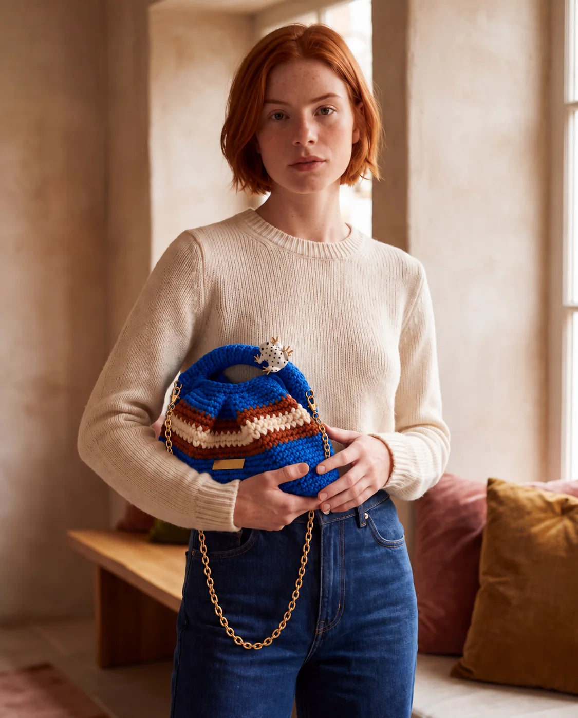 Model in an ecru jumper and jeans holding Clutch Urbana Blue by The Labbo, with its gold chain, beside a window.