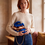 Model in an ecru jumper and jeans holding Clutch Urbana Blue by The Labbo, with its gold chain, beside a window.