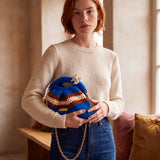 Model in an ecru jumper and jeans holding Clutch Urbana Blue by The Labbo, with its gold chain, beside a window.