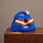 Clutch Urbana Blue by The Labbo, with terracotta and ecru stripes, a gold chain and an enamelled frog, on a wooden ledge.