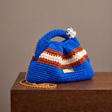 Clutch Urbana Blue by The Labbo, with terracotta and ecru stripes, a gold chain and an enamelled frog, on a wooden ledge.