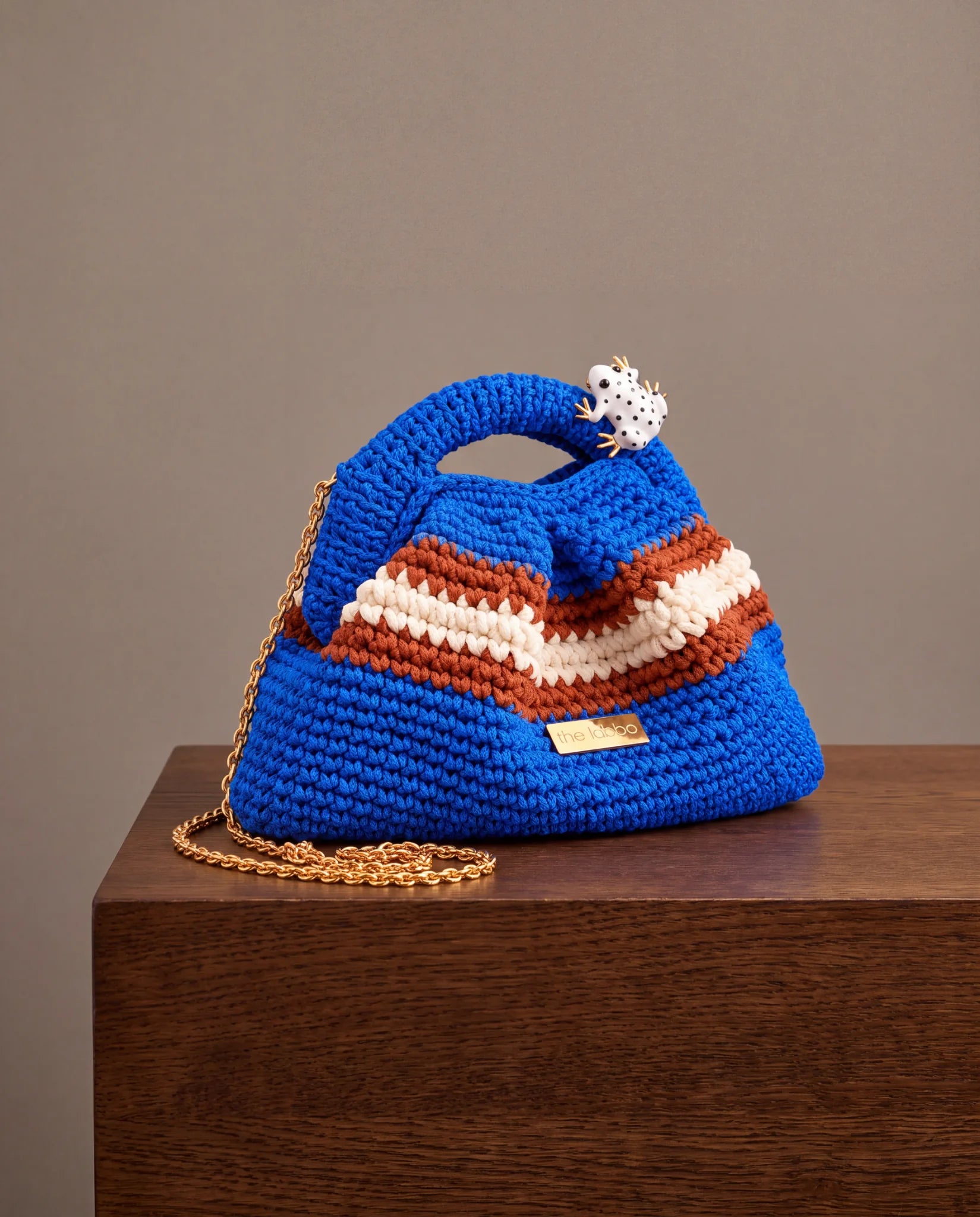 Clutch Urbana Azul de The Labbo, con franjas terracota y crudo, cadena dorada y ranita esmaltada, sobre una repisa de madera.