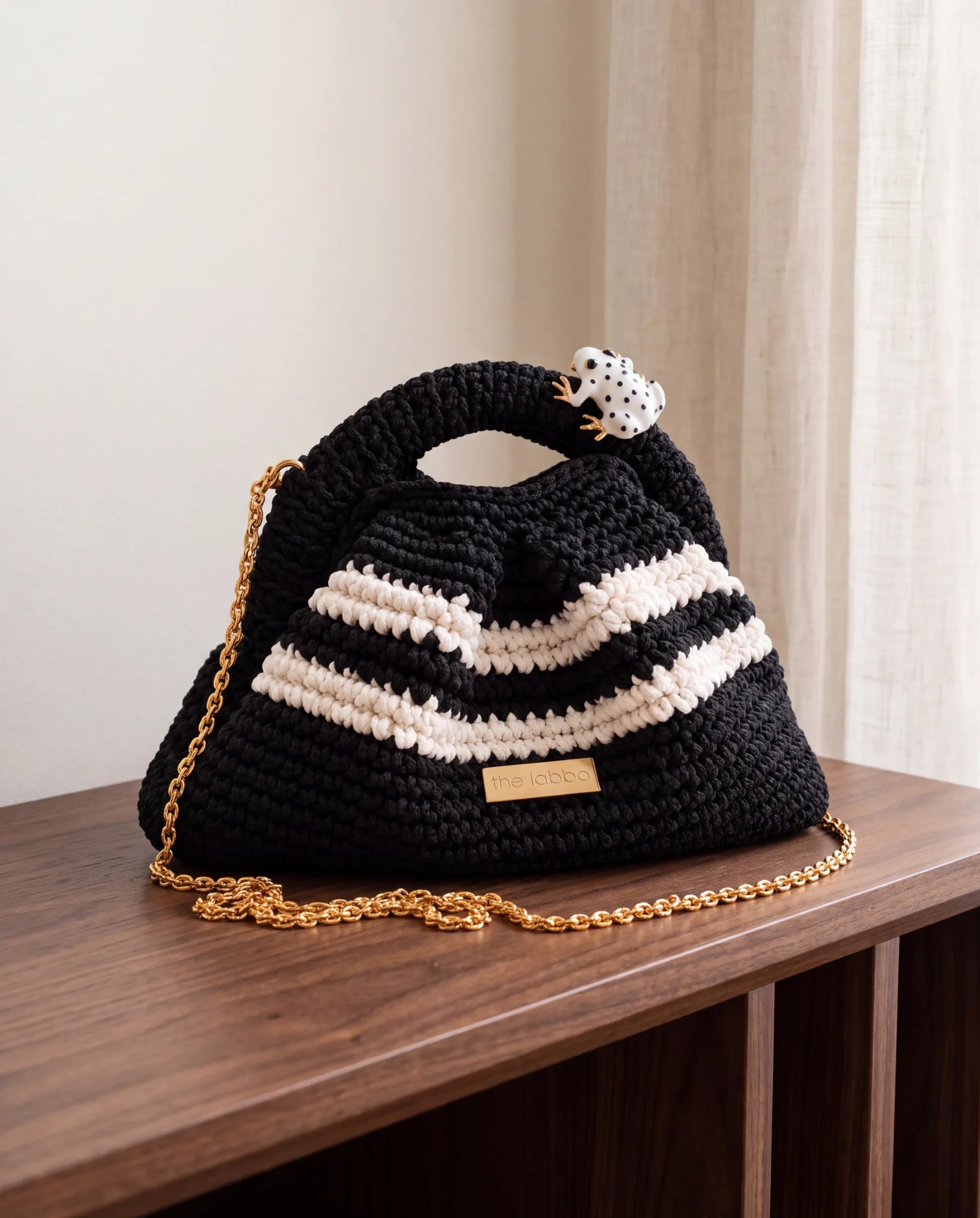 Clutch Urbana Negro y Blanco de The Labbo con cadena dorada y ranita esmaltada, sobre una consola de madera junto a una cortina.