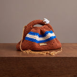 Clutch Urbana Terracota de The Labbo, con franjas azul y crudo, cadena dorada y ranita esmaltada, sobre una repisa de madera.