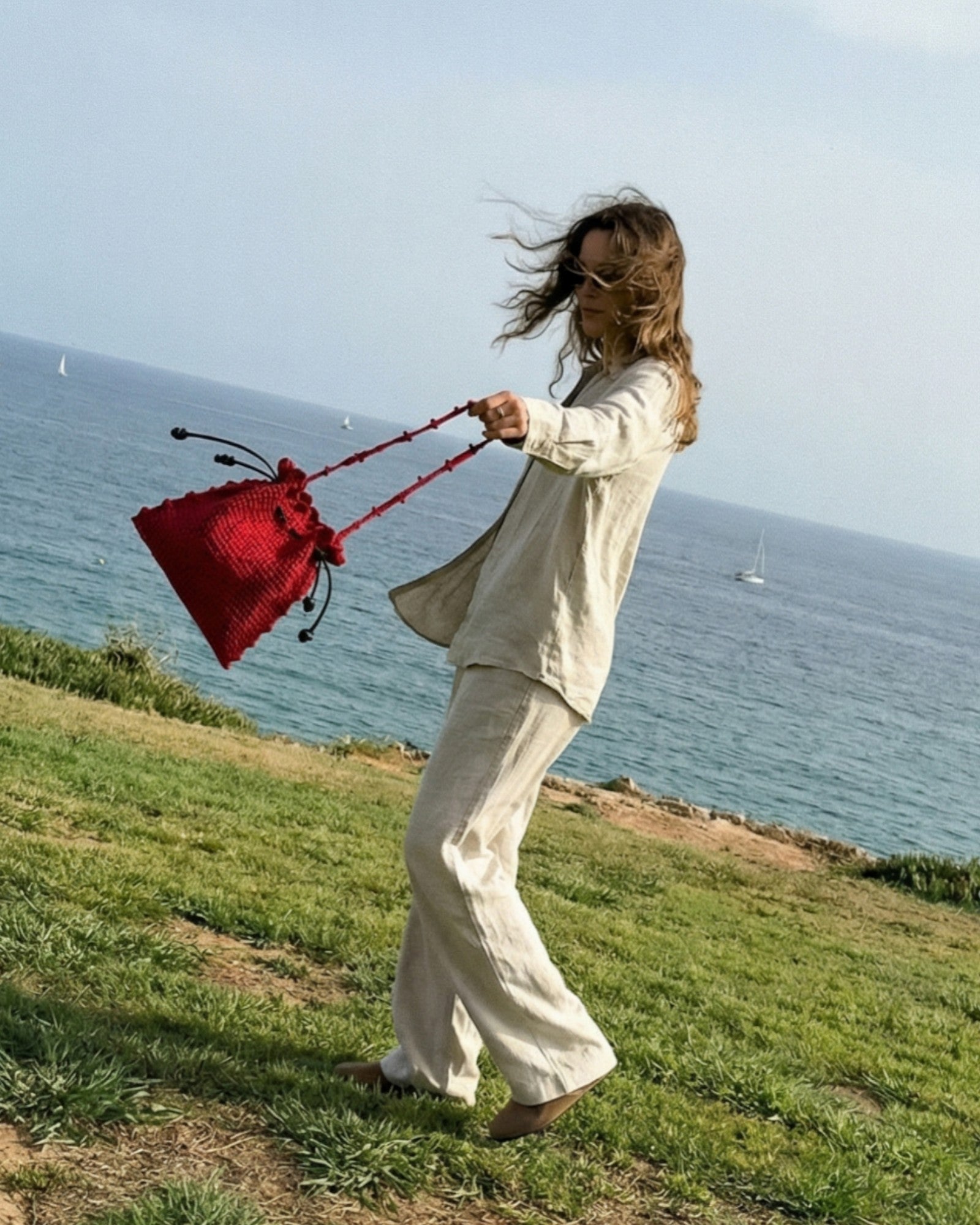 Bolso rojo hecho a mano en crochet con silueta recogida, sujetado por asa tejida a mano en un paisaje con viento